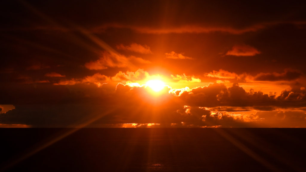 A photo of a fiery orange Hawaiian sunset with fluffy clouds partially obscuring the sun.