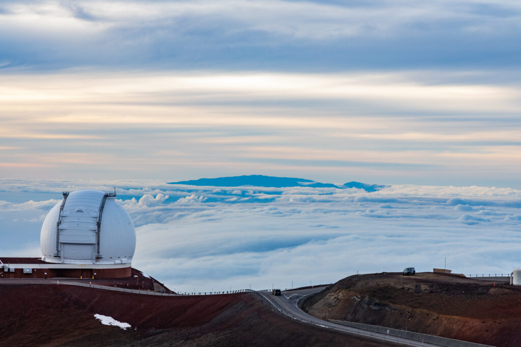 A photorealistic image from the summit of Mauna Kea looking out at the horizon, with a faint outline of Maui in the distance and a clear blue sky.