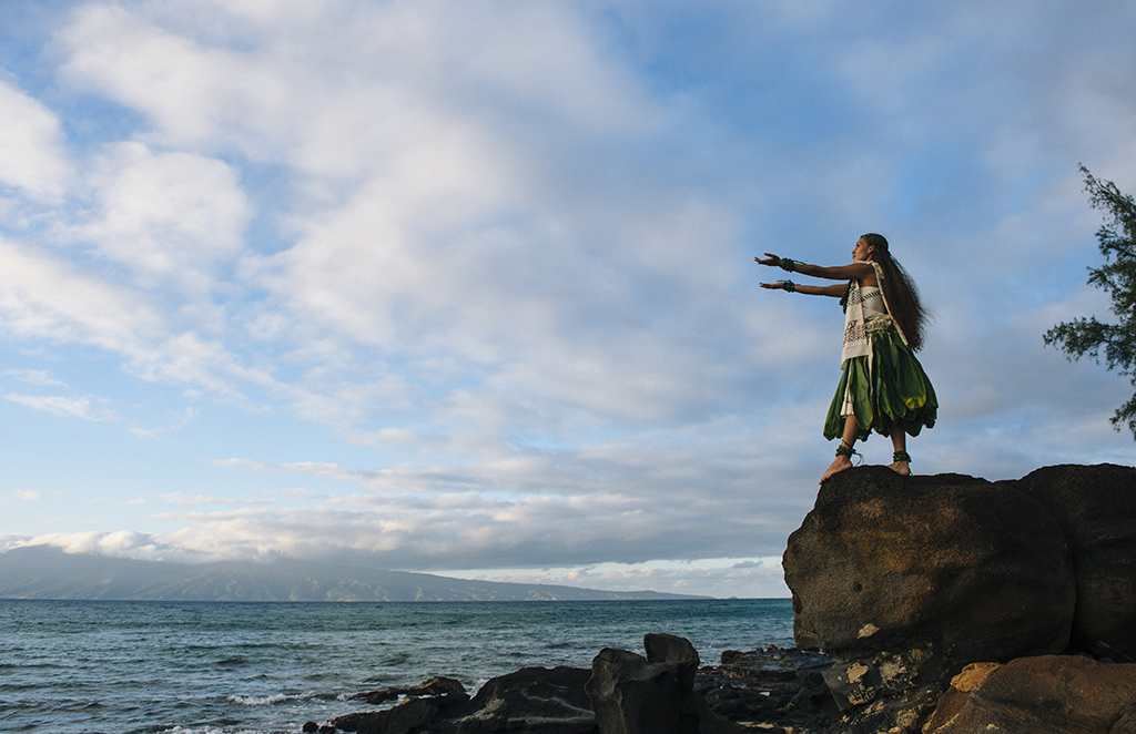 Woman hula dancing on top of coastal rocks wearing traditional c